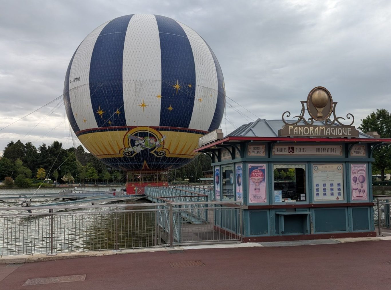 A large hot air balloon over a park near a kiosk by a water body.