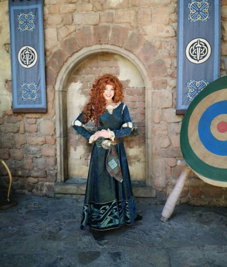 A woman in a long green dress with red curly hair, standing near a stone archway and target.