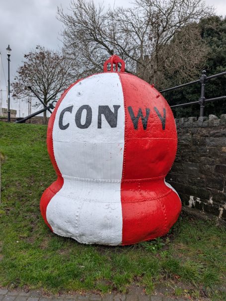 Large red and white buoy marked with the word "CONWY" near a coastal area.