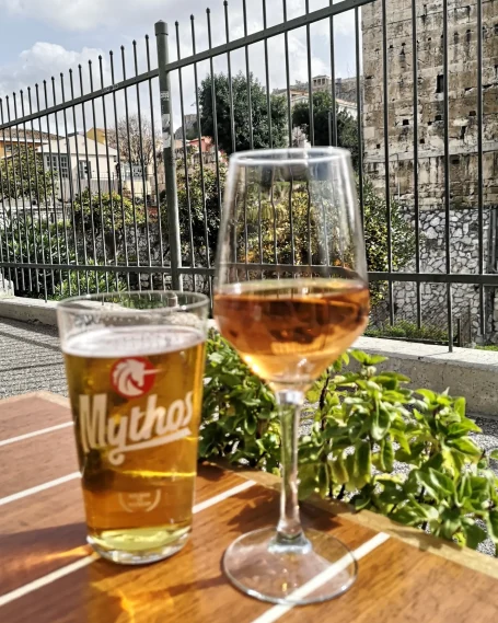 Two drinks on a wooden table: a pint of beer and a glass of rosé wine, with a view of greenery and the ancient city of Athens.