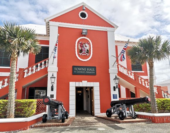 Historic St George's Town Hall building with a red facade, featuring palm trees and cannons at the entrance. Located in Bermuda.