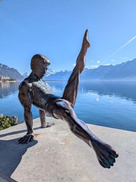 Statue Li Ning, a bronze sculpture of a dancer posed on the edge of Lake Geneva with mountains in the background.