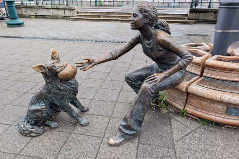 Girl With Her Dog Statue: Bronze statue of a girl feeding a dog in a public square in Budapest.