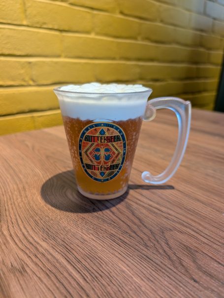 A glass of Butterbeer, placed on a wooden table against a yellow wall.