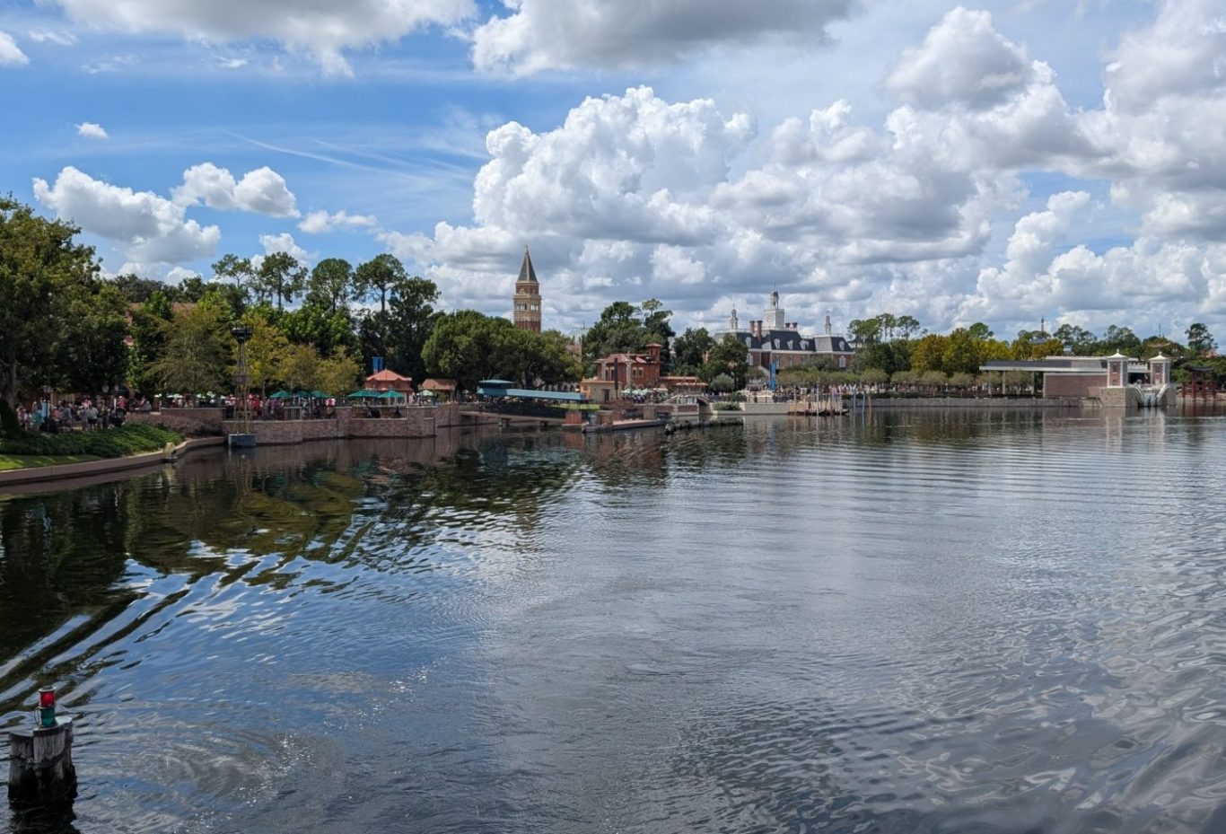View of a calm lake surrounded by trees and distant buildings under a cloudy sky.