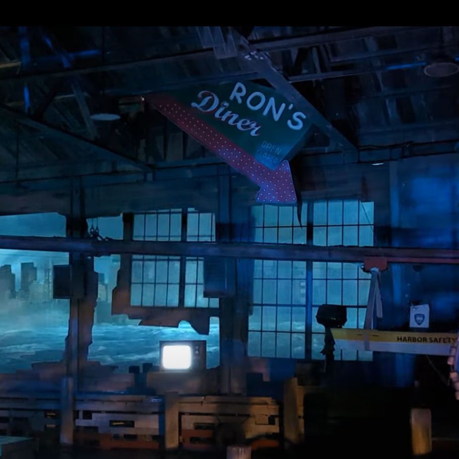 Interior of a dimly lit diner with a sign pointing to "Ron’s Diner" amidst blue tones.