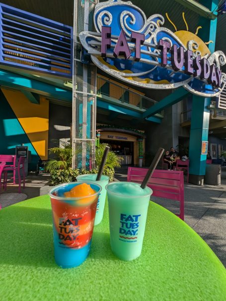 Two vibrant frozen drinks sit on a bright green table outside a Fat Tuesday venue.
