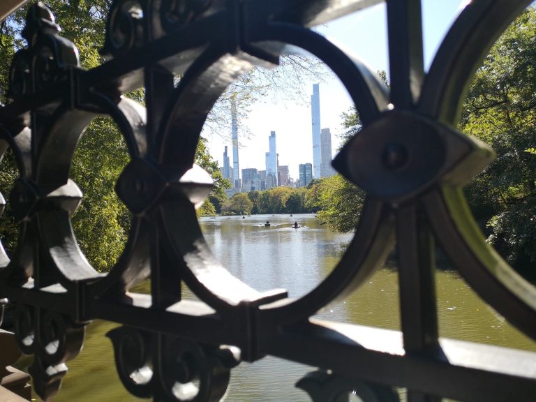 View of a serene river framed by decorative ironwork, with New York city skyscrapers in the background.