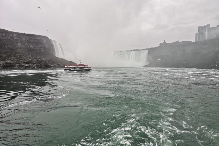 Boat navigating near the misty waterfalls of Niagara Falls with rocky cliffs and a cloudy sky.