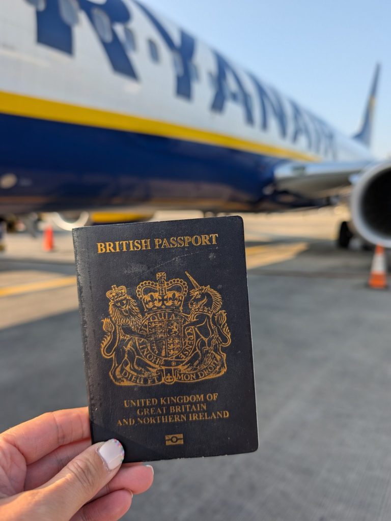 A hand holding a British passport in front of a Ryanair airplane.