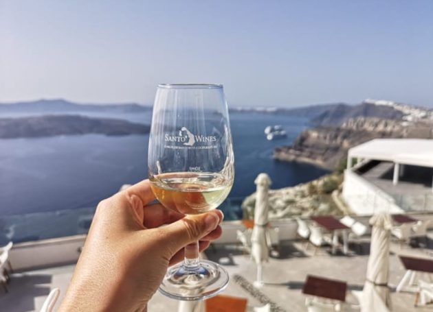 A hand holding a Santos wine glass with a view of the sea and cliffs in Santorini.