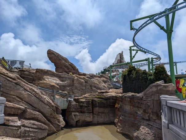 Rocky landscape with water at the base and roller coaster tracks above under a blue sky.