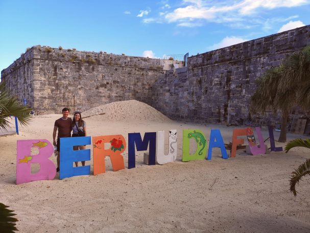 I&B's Founder & Husband standing beside colourful letters spelling "BERMUDA" on a sandy beach with ruins in the background.