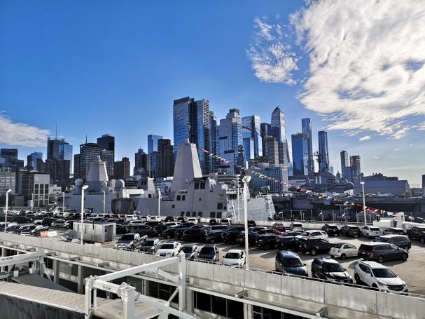 New York City skyline of tall buildings under a blue sky, with boats and cars in the foreground. The view from Manhattan Cruise Terminal.