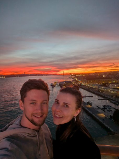 I&B's Founder & Husband posing on a cruise ship with the sunset over the city of Lisbon in the background.