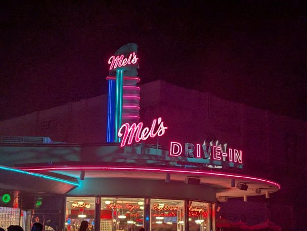 Neon-lit retro diner sign at night, featuring "Mel's" in vibrant colours.