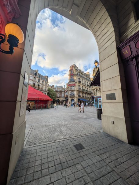 View of a square through an archway, featuring historic buildings and cloudy sky.