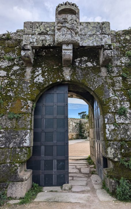 Stone archway with a metal door, surrounded by moss and leading to an outdoor landscape at Castelo do Castro in Vigo Spain.