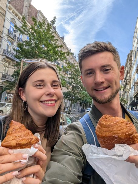 I&B's Founder & Husband posing on a street in Brussels with croissants, which a cloudy blue sky in the background.