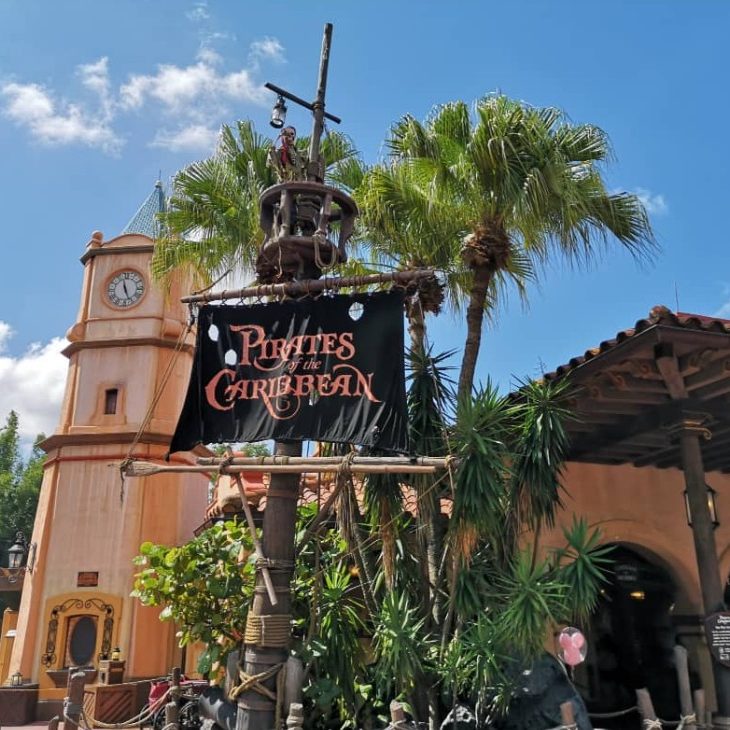 Entrance to a themed attraction with a pirate banner, palm trees, and a clock tower.