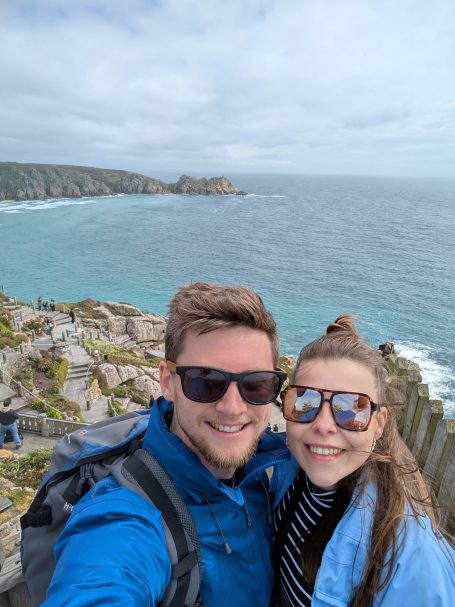 I&B's Founder & Husband posing with the clear blue waters and Minack Theatre in the background