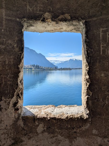 View through a stone wall at Chillon Castle showing the serene Lake Geneva and mountains under a clear blue sky.
