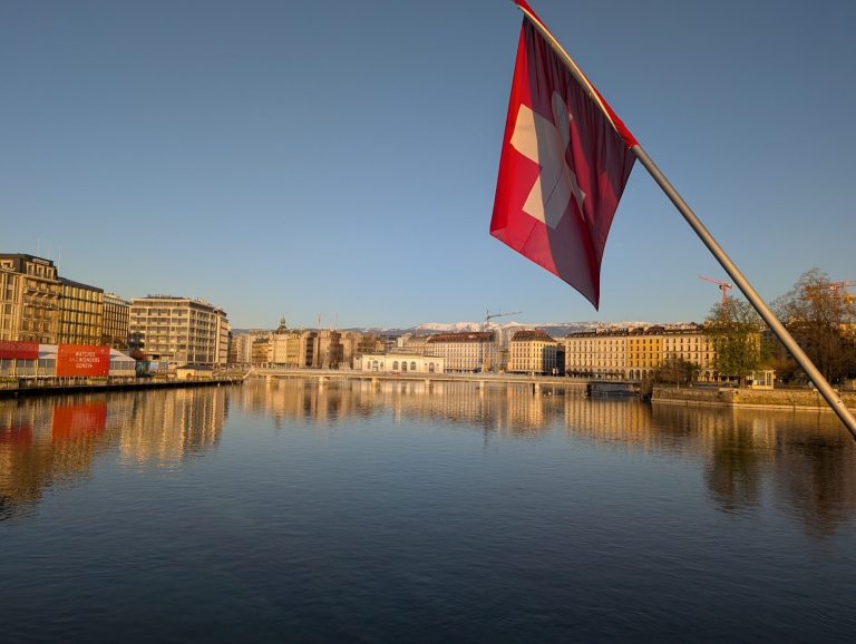 Swiss flag in the foreground with a tranquil river and buildings reflected in the background.