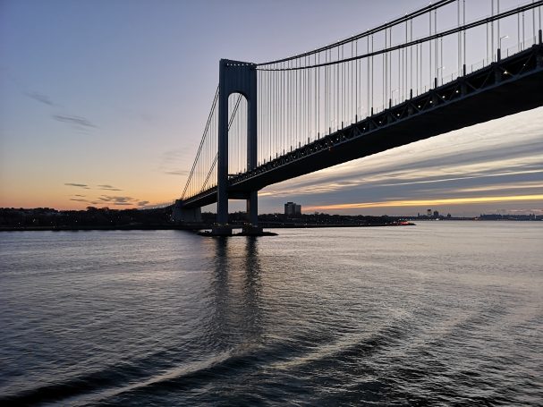Sunset view of a suspension bridge over calm waters with a city skyline in the background.
