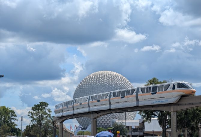 The Disney monorail passes by the iconic geodesic sphere of Epcot under a cloudy sky.
