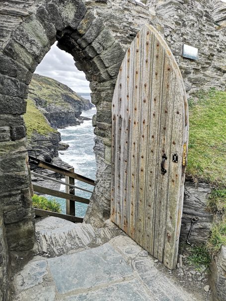 Weathered wooden door set in a stone archway, leading to a coastal view.