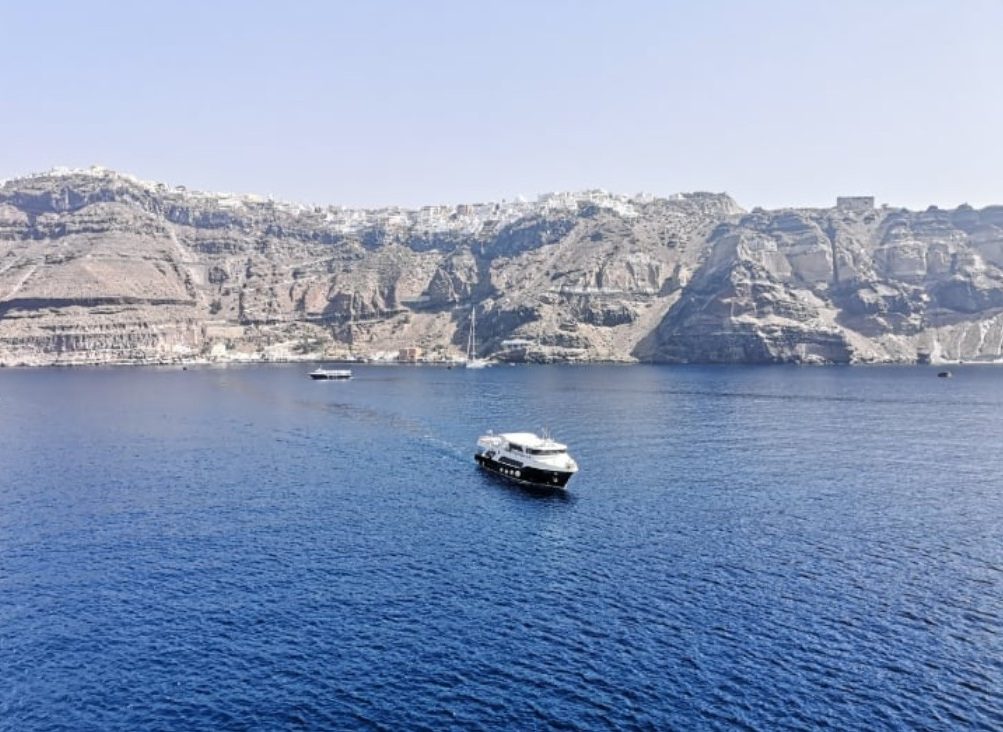 A yacht sailing on calm blue waters near rocky cliffs. Views of Santorini from a cruise ship.