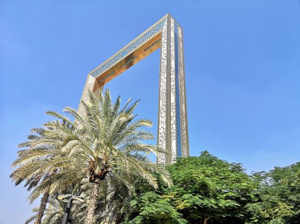 A tall, golden frame-shaped structure, known as the Dubai Frame surrounded by palm trees against a blue sky.