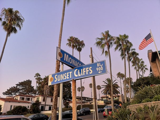Street signs for Sunset Cliffs and another road, with palm trees and a flag in the background.