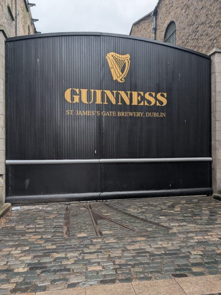 Black wooden gate with golden Guinness logo and text, set against cobblestone path.