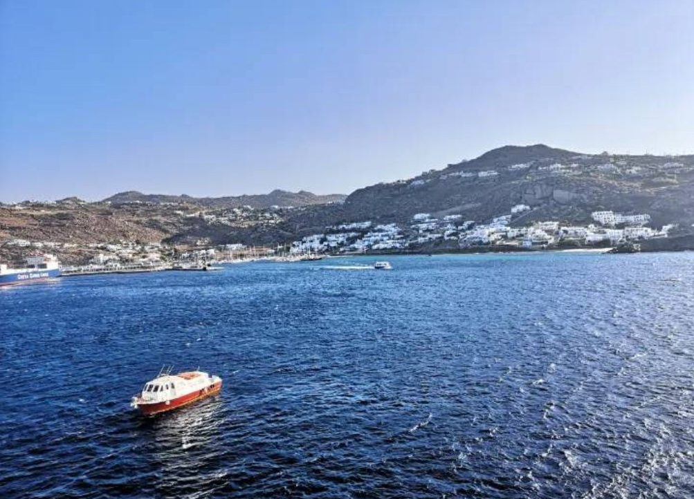 A small boat on calm blue waters with hills and white buildings in the background. View of Mykonos from a cruise ship.
