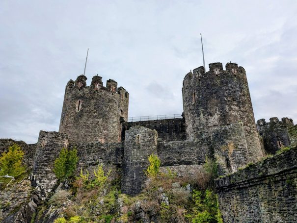 Stone castle with two towers, surrounded by greenery under a cloudy sky.