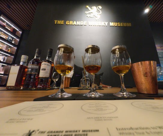 Three tasting glasses of whisky, with bottles and a menu in the background, located in The Grande Whisky Museum in Singapore.