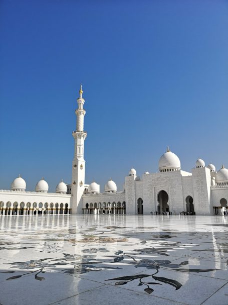 White mosque with a tall minaret against a clear blue sky. Reflective surface in foreground.