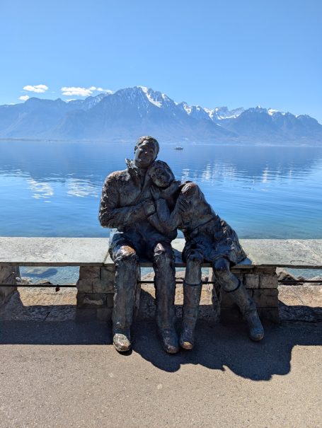 Two bronze statues of a couple seated by Lake Geneva with mountains in the background.