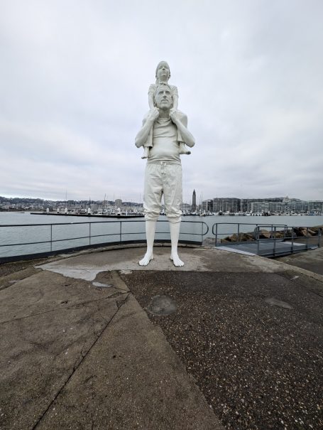 Jusqu'au Bout du Monde, a large white statue of a man holding a child, overlooking a body of water.