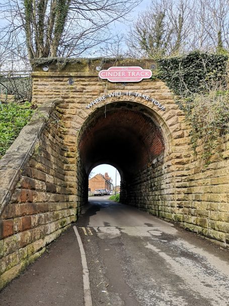 Stone archway tunnel with a sign indicating the start of The Cinder Track, leading to a road and houses beyond, located in Whitby.