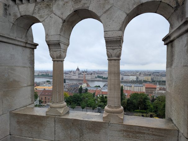 View of the Budapest city skyline through arched stone columns at Fisherman's Bastion on a cloudy day.