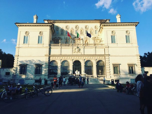 Historic Galleria Borghese building with flags, featuring a grand entrance and people gathered outside, located in Villa Borghese in Rome.
