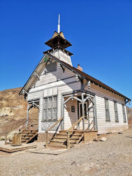 Old wooden schoolhouse with a bell tower set against a blue sky and arid landscape, located at Calico Ghost Town in California.