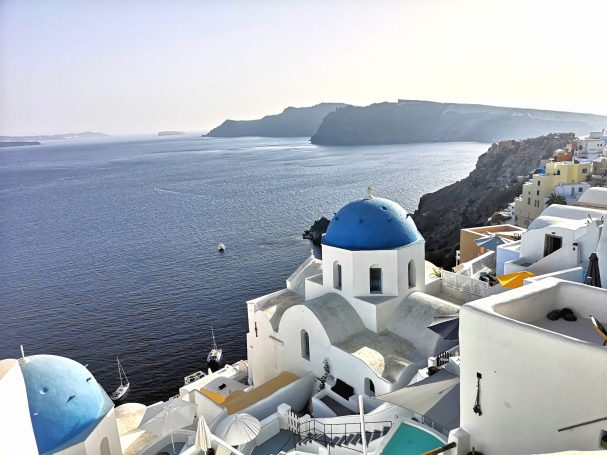 White buildings with blue domes overlooking a calm sea and mountainous coastline.