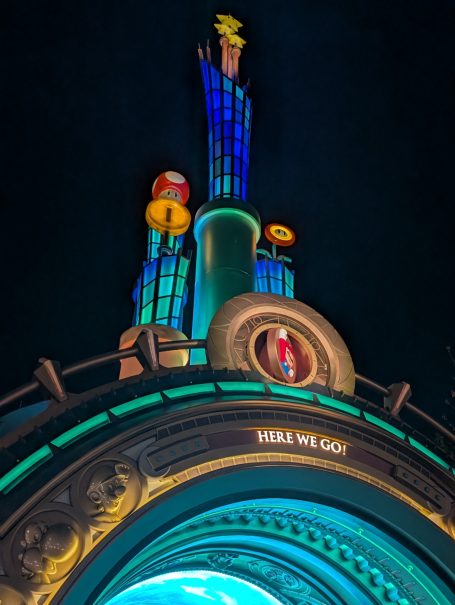 Brightly lit tower with colourful icons and a clock displaying "Here We Go!" at night.