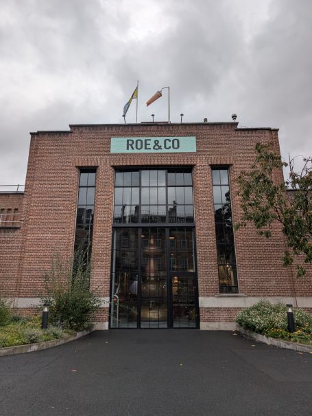 Brick building with large glass doors, featuring flags and "ROE & CO" sign above.