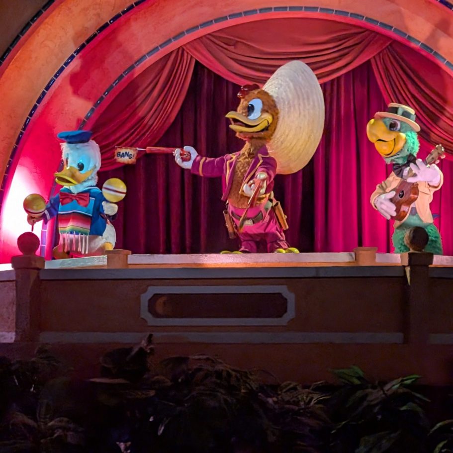 Three animated ducks in festive attire perform on a stage with red curtains.