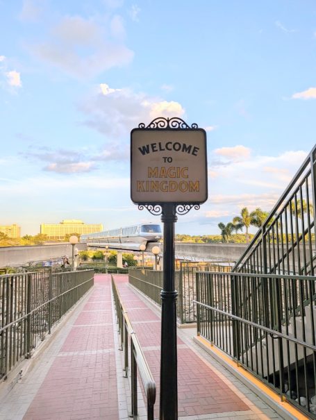 Welcome to Magic Kingdom sign on a pathway leading towards a river with blue sky above.