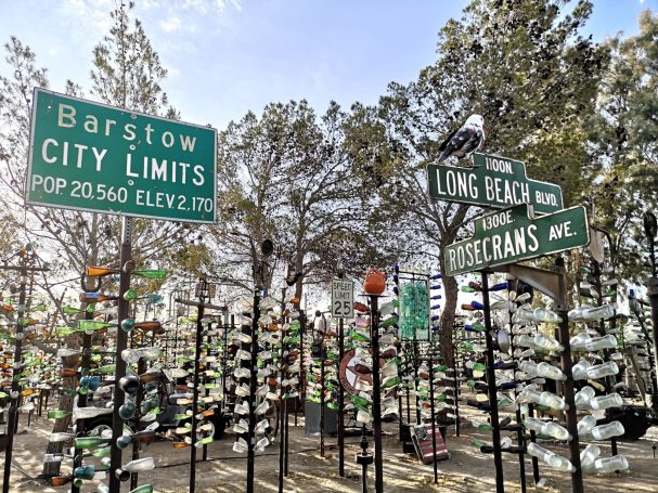 Colourful glass bottles arranged on poles, to look like trees at Elmer’s Bottle Tree Ranch, with Barstow and Long Beach street signs.